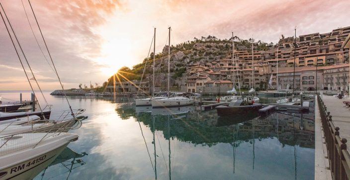 The sun setting behind a rocky hill in Portopiccolo, Italy with sailboats and crystal clear water in the foreground.