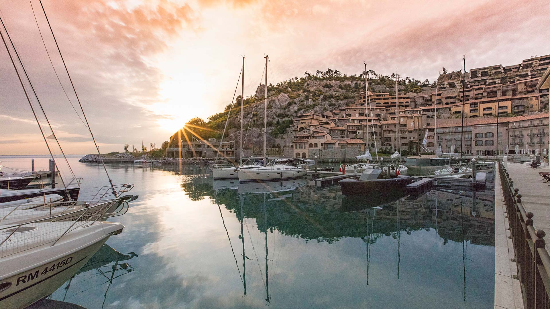 The sun setting behind a rocky hill in Portopiccolo, Italy with sailboats and crystal clear water in the foreground.