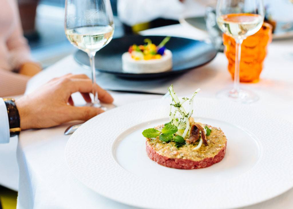 person holding glass of drink on table beside plate of dish