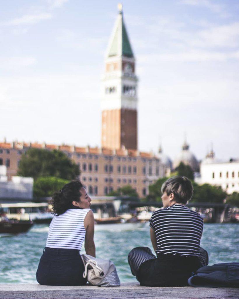 selective focus photo of two women talking while sitting in front body of water at daytime