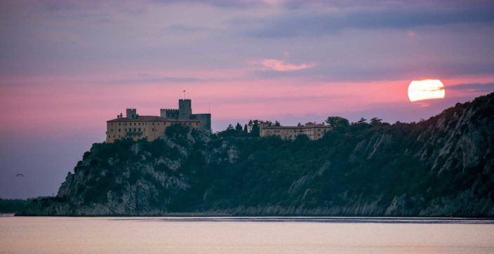 a fourteenth century castle on a cliffside by the water with the sun setting in the background