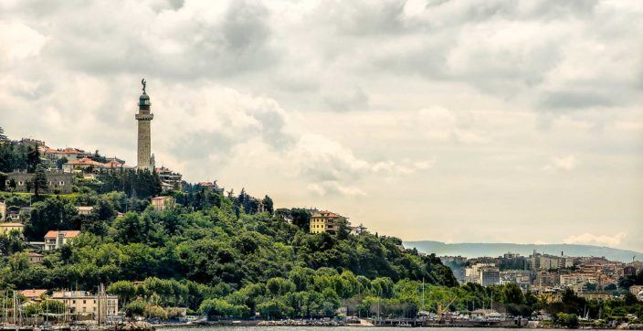 a tall and ornate lighthouse standing above a tree-covered Italian city slope
